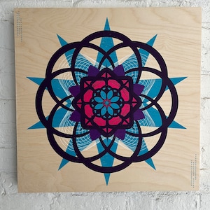 May include: A geometric mandala design with a blue, purple, and pink color scheme. The design is printed on a wood panel.