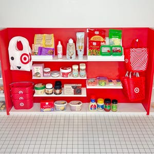 May include: A red and white miniature grocery store display with shelves stocked with various food and household items. The shelves are filled with bags of snacks, jars of spices, bottles of cleaning supplies, and other grocery items. There are also two red shopping bags with the Target logo hanging on the side of the display.
