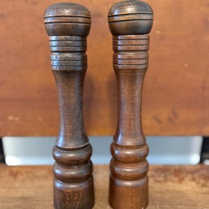 May include: Two vintage wooden salt and pepper grinders. The grinders are made of dark brown wood with a rounded top and a ridged design. One grinder has a brass-colored top. The grinders are standing upright on a wooden surface.