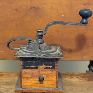 May include: Antique wooden coffee grinder with a black metal crank and a drawer. The grinder has a label that reads "A.C. Mill".