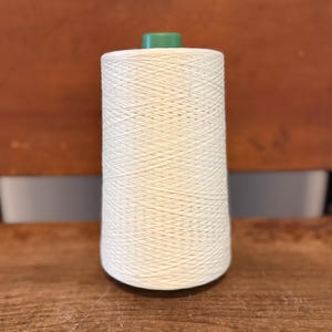 May include: A large spool of off-white thread with a green cap. The thread is tightly wound around a conical core. The spool is set on a wooden surface with a brown background.