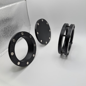 May include: Three black, circular objects with embedded magnets. One is a ring, another a flat disc, and the third is a ring held together by screws. The objects are on a white surface, with a silver backdrop.