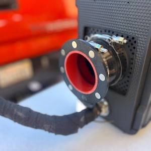 May include: Close-up of a black camera with a red and black exhaust port. The camera has a textured black exterior and a silver metal part on the side.