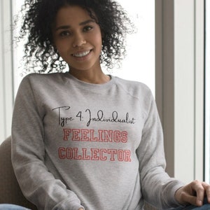 Puede incluir: Una mujer lleva una sudadera gris con el texto "Type 4 Individualist FEELINGS COLLECTOR" en letras negras y rojas. Está sentada en una silla con las piernas cruzadas y sonriendo a la cámara.