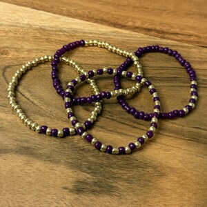 May include: Four beaded bracelets in gold and purple. The bracelets are made of small, round beads strung together. The bracelets are resting on a wooden surface.