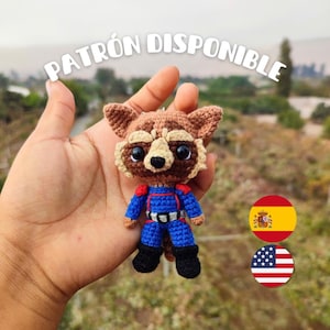 Könnte beinhalten: Ein gehäkeltes Rocket Raccoon Amigurumi-Spielzeug in einem blauen und roten Overall. Das Spielzeug wird in einer Hand gehalten, über dem der Text "PATRÓN DISPONIBLE" steht. Die spanische und amerikanische Flagge sind im Hintergrund zu sehen.