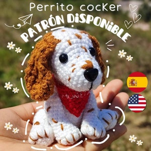 Op de afbeelding: Een gehaakte witte en bruine cocker spaniel puppy die een rode bandana draagt met het woord "MILWEITA" erop. De puppy zit in een hand met een witte achtergrond. De tekst "PATRÓN DISPONIBLE" is in wit op een zwarte achtergrond boven de puppy geschreven. De tekst "Perrito cocker" is in wit op een zwarte achtergrond boven de tekst "PATRÓN DISPONIBLE" geschreven. De tekst "@gatos_lanudos" is in wit op een zwarte achtergrond onder de puppy geschreven.