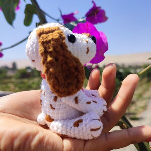 May include: A small crocheted dog toy with brown and white fur. The dog is sitting in a hand with its head tilted up, looking at a purple flower.