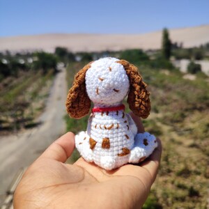 May include: A white crocheted dog with brown ears and spots. The dog is sitting on a hand with a blurry background of a desert landscape.