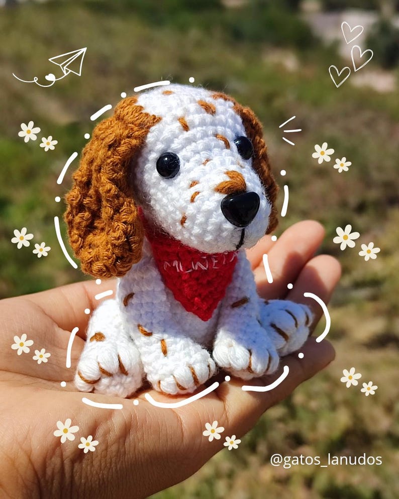 May include: A crocheted white and brown dog with a red bandana. The dog is sitting in a hand and has a black nose and brown eyes.