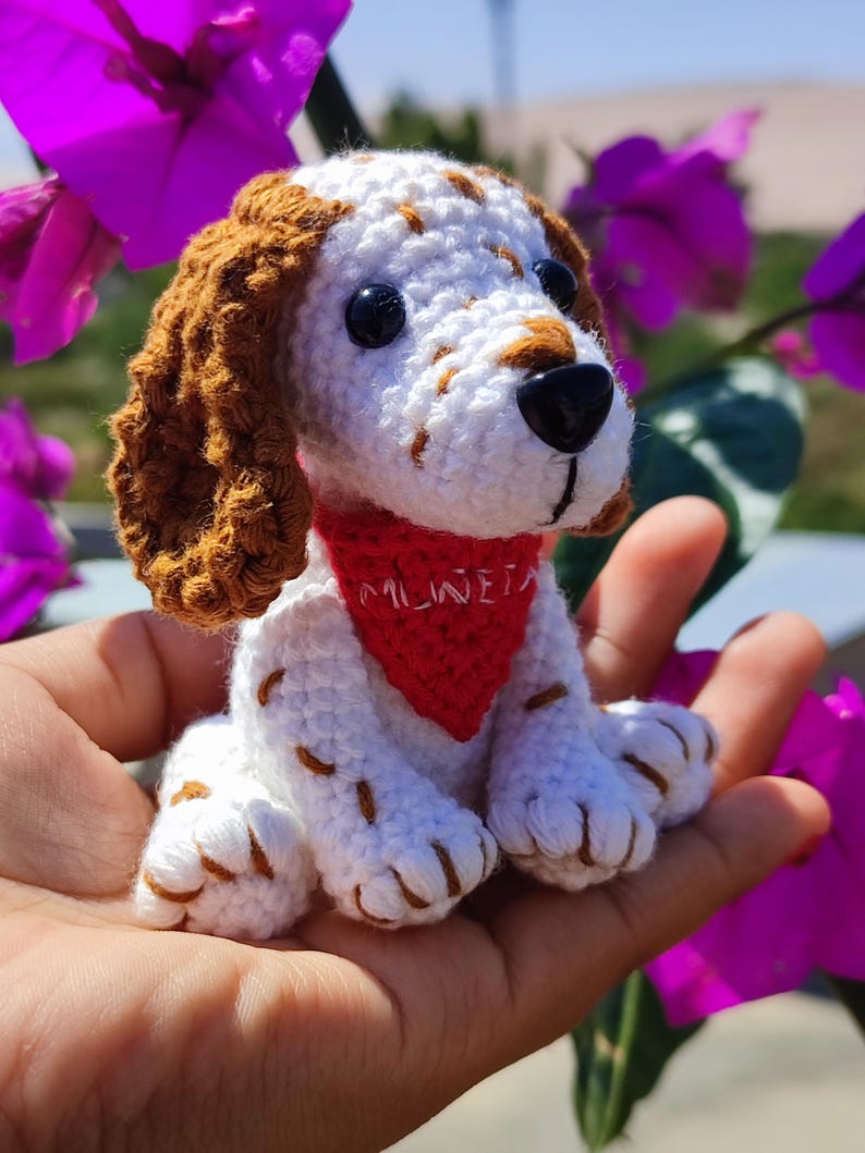 May include: A crocheted white and brown dog with a red bandana that says "MILWE" sitting in a hand. The dog has black eyes and a black nose.