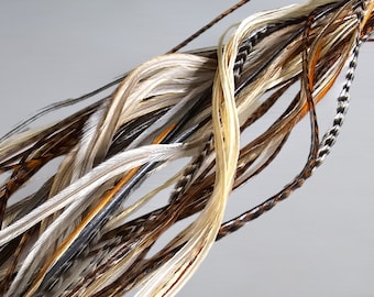 Puede incluir: Un manojo de plumas largas y delgadas en varios tonos de marrón, blanco y negro. Las plumas están dispuestas en un patrón suelto y fluido, creando una textura suave y delicada.
