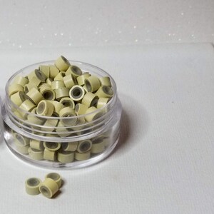 Puede incluir: Un recipiente de pl&aacute;stico transparente lleno de peque&ntilde;as cuentas de silicona beige claro. Las cuentas se utilizan para extensiones de cabello de micro-loop.