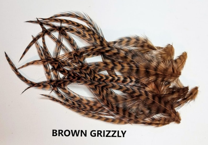May include: A bunch of brown grizzly feathers with black barring. The feathers are arranged in a loose pile on a white background. The text "BROWN GRIZZLY" is printed below the feathers.