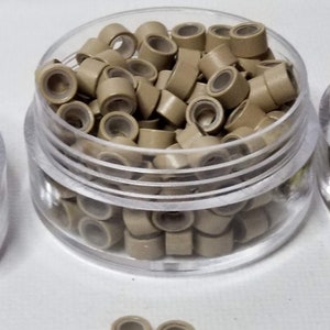 Puede incluir: Un recipiente de pl&aacute;stico transparente lleno de peque&ntilde;os microanillos marrones claros para extensiones de cabello.