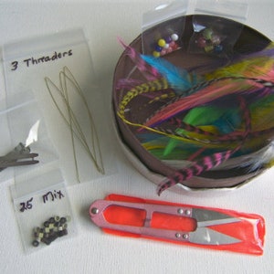 Op de afbeelding: Assortiment aan knutselbenodigdheden voor het maken van verenhaarverlengingen, waaronder 3 draadjes, 2 clips, 25 mix kralen, kleurrijke veren en een klein schaartje.