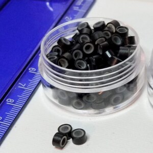 Puede incluir: Un recipiente de pl&aacute;stico transparente lleno de microanillos negros para extensiones de cabello. Tambi&eacute;n hay tres microanillos sobre una superficie blanca.
