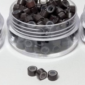 Puede incluir: Un recipiente de pl&aacute;stico transparente lleno de peque&ntilde;os microanillos marrones y plateados para extensiones de cabello.