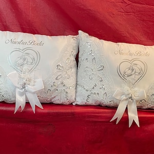 May include: Two white wedding ring pillows with intricate lace and sequin detailing. Each pillow features the text "Nuestra Boda" above a heart-shaped design with interlocking rings. White ribbon bows add a decorative touch.
