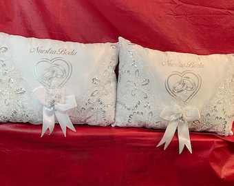 May include: Two white wedding ring pillows with intricate lace and sequin detailing. Each pillow features the text "Nuestra Boda" above a heart-shaped design with interlocking rings. White ribbon bows add a decorative touch.