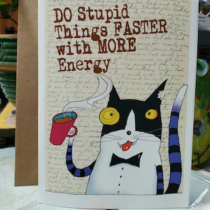Può includere: Biglietto d'auguri con un gatto dei cartoni animati che tiene una tazza di caffè. Il gatto è bianco e nero con un papillon. Il testo della carta recita "Drink Coffee, Do Stupid Things Faster with More Energy."