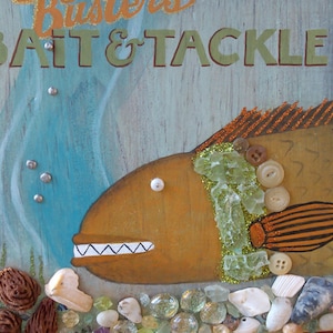 Puede incluir: Un letrero decorativo con un pez naranja estilizado con una sonrisa amplia y dentada. El pez está adornado con botones y purpurina, sobre un fondo azul y verde. El letrero dice "Busters Bait & Tackle" en letras naranjas y verdes, con elementos decorativos de conchas y vidrio.