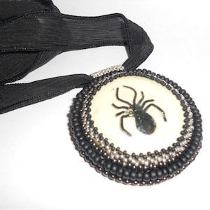 Puede incluir: Un collar con colgante de araña negra con una cinta negra. El colgante tiene un fondo color crema con un diseño de araña negra. El colgante está rodeado de cuentas negras y plateadas.