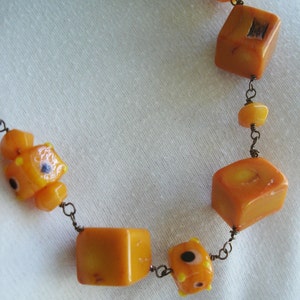May include: A necklace featuring orange, cube-shaped beads and smaller, rounded beads. The beads are strung together with a dark metal chain. Some beads have small, dark accents.