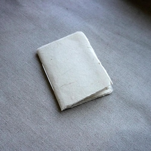 May include: A small, off-white handmade paper notebook with a rough, textured surface. The pages are folded and bound, creating a compact, rectangular shape. The notebook is set against a gray fabric background.