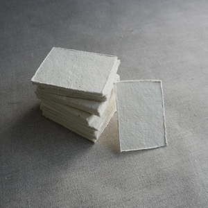 Op de afbeelding: Een stapel vierkante, gebroken witte handgemaakte papieren kaarten met ruwe, getextureerde randen. Eén kaart ligt apart, naast de stapel. De kaarten liggen op een grijze, getextureerde ondergrond.