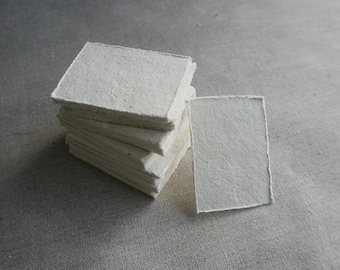 May include: A stack of square, off-white handmade paper cards with rough, textured edges. One card is separate, lying next to the stack. The cards are on a gray, textured surface.