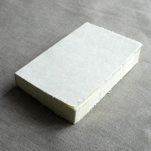 Puede incluir: Una pila de hojas de papel rectangulares de color blanquecino con bordes ásperos y recortados. El papel tiene una superficie texturizada y está cuidadosamente apilado sobre un fondo de tela gris. El papel parece hecho a mano.