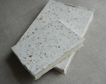 May include: Two handmade paper journals, one large and one small, are stacked on a neutral-toned surface. The covers are a speckled off-white with visible flecks of brown and tan. The pages are a creamy white.
