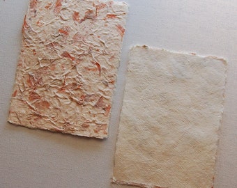 May include: Two sheets of handmade paper, one with a textured surface and orange flecks, the other plain white.