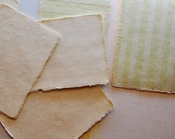 以下が含まれることがあります： 不揃いの縁取りのある手作りの紙の束。一番上の紙は薄い緑色で白い縞模様。他の紙はクリーム色です。