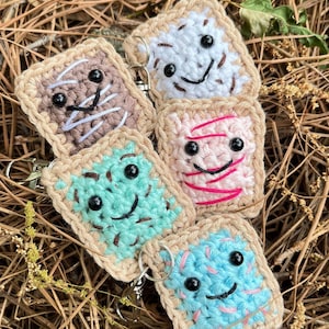 May include: Four crocheted keychains shaped like toast with different colored fillings. The fillings are white, pink, blue, and brown. Each keychain has a smiling face with black eyes.