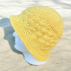 May include: A bright yellow crochet bucket hat featuring a textured starburst pattern throughout the crown and sides. The hat has a short, downward-sloping brim and is displayed on a white mannequin head to show its rounded shape and structure.
