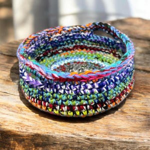 May include: A handmade, colorful crochet basket with a round shape and a handle. The basket features a vibrant mix of blue, green, red, orange, and purple yarns, creating a textured and eye-catching design. The basket is sitting on a wooden surface.