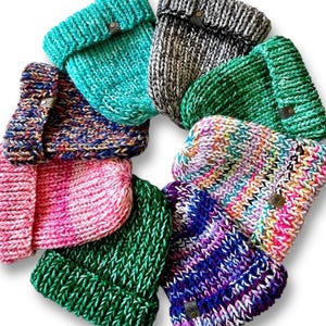 May include: A circular arrangement of nine chunky knit beanies in various colors including turquoise, grey, green, pink, and multi-colored yarns. Each hat features a thick folded brim and a small brown leather-like tag attached to the side.