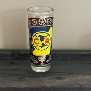 May include: A clear glass shot glass with a silver metal band featuring a soccer ball design. The band also includes a blue and yellow logo with the letters "C" and "A" and a trophy.