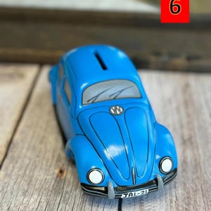 May include: Blue ceramic piggy bank shaped like a Volkswagen Beetle. The car has a slot on the roof for coins and a license plate that reads "JAL-21".