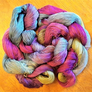 May include: A skein of hand-dyed yarn in a variety of colors, including teal, pink, purple, and gold. The yarn is arranged in a braided pattern, with the strands of yarn visible. The yarn is on a wooden surface.