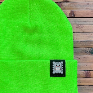 May include: Vibrant neon green beanie hat with a turned-up brim. A small, square patch with a cartoon face is sewn on the brim. The text "RAPSCALLION" and "RAPSCALLION.CA" are printed in white at the bottom.