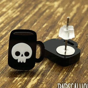 Peut inclure: Boucles d'oreilles en acrylique noir représentant une tasse à café avec un motif de crâne blanc. Les boucles d'oreilles ont une tige et un fermoir pour oreilles percées. Le nom de la marque "RAPSCALLION" est imprimé en blanc en bas.