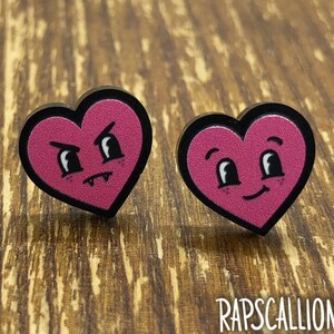 May include: A pair of heart-shaped earrings. Each earring has a black border and a pink heart design. One has an angry face, the other a smiling face. The word "RAPSCALLION" is printed in white at the bottom.
