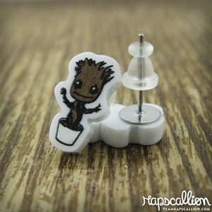 May include: A pair of white stud earrings with a cartoon Groot design. The Groot image is brown and black, in a white pot. The earrings have a silver post and a clear backing. The background is a wooden surface.
