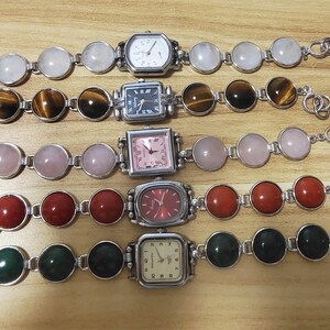 May include: Five silver-toned bracelet watches with various gemstone and watch face designs. The gemstones include white, brown, pink, red, and green. The watch faces are square and rectangular, with different colored dials and hands.