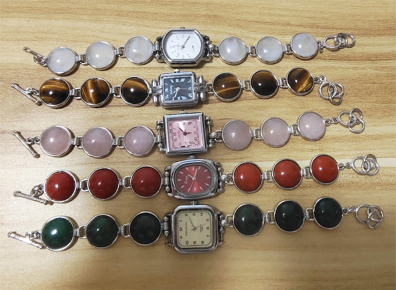 May include: Five silver-toned bracelet watches with various gemstone and watch face designs. The gemstones include white, brown, pink, red, and green. The watch faces are square and rectangular, with different colored dials and hands.