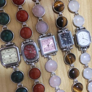 May include: Collection of gemstone watches with various colored stones and watch faces. The watches feature round and square faces, with colors including red, pink, green, and tiger's eye. The watch bands are made of silver-toned metal.