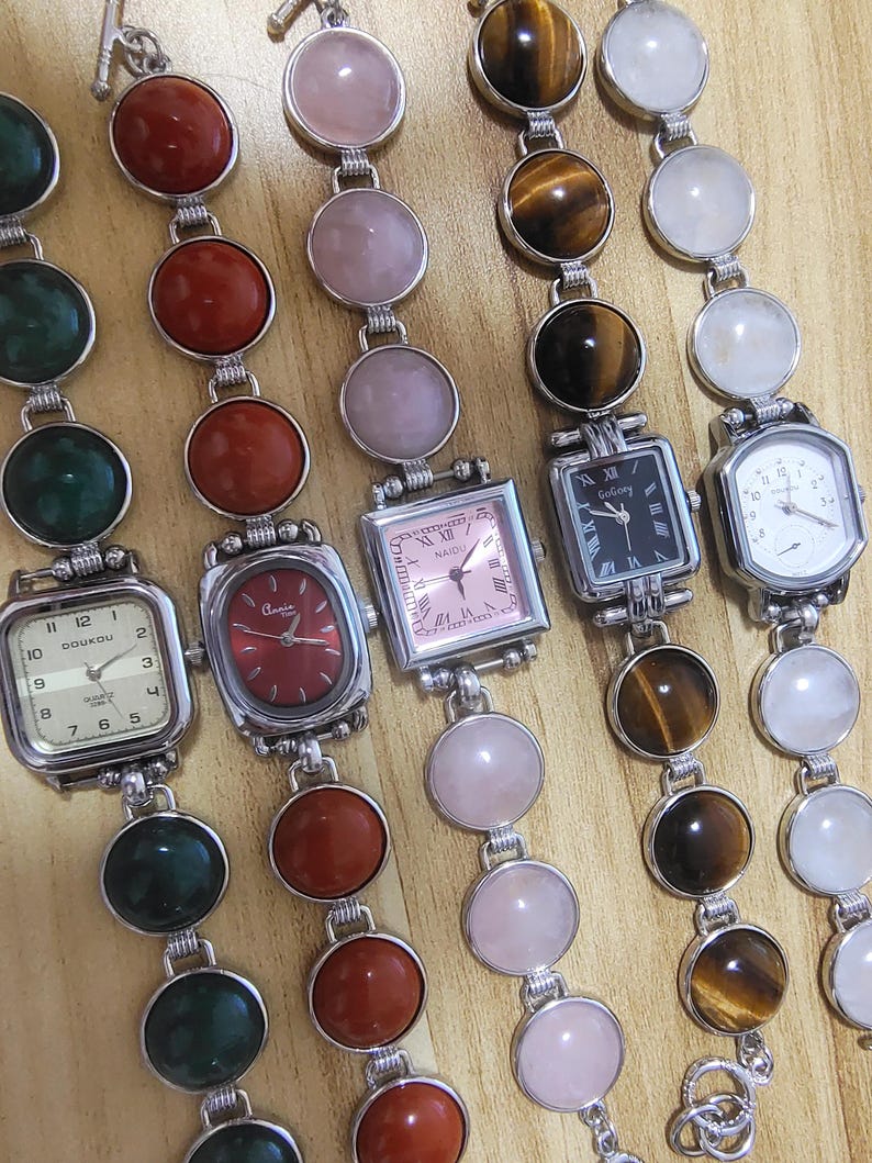 May include: Collection of gemstone watches with various colored stones and watch faces. The watches feature round and square faces, with colors including red, pink, green, and tiger's eye. The watch bands are made of silver-toned metal.